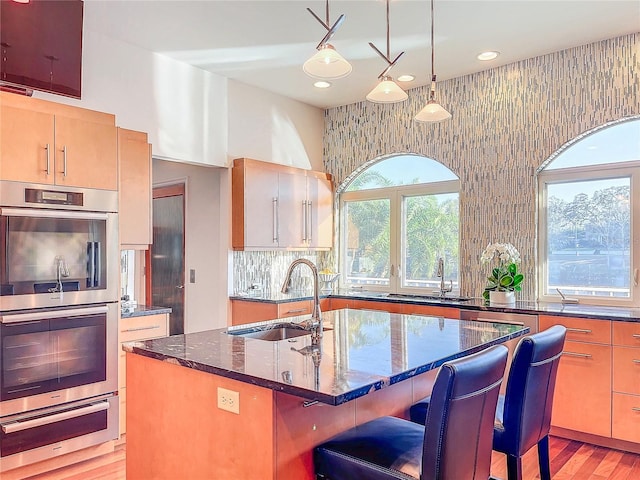 kitchen featuring pendant lighting, a kitchen bar, dark stone counters, a kitchen island with sink, and stainless steel double oven