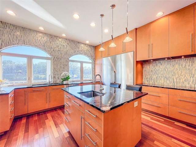 kitchen featuring sink, hanging light fixtures, a center island with sink, stainless steel fridge, and dark stone counters