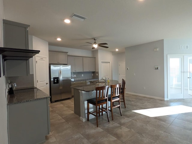 kitchen with a kitchen bar, gray cabinetry, stainless steel fridge, a kitchen island with sink, and backsplash