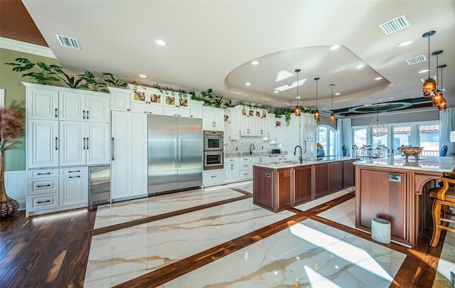 kitchen featuring a raised ceiling, appliances with stainless steel finishes, a kitchen island with sink, and decorative light fixtures