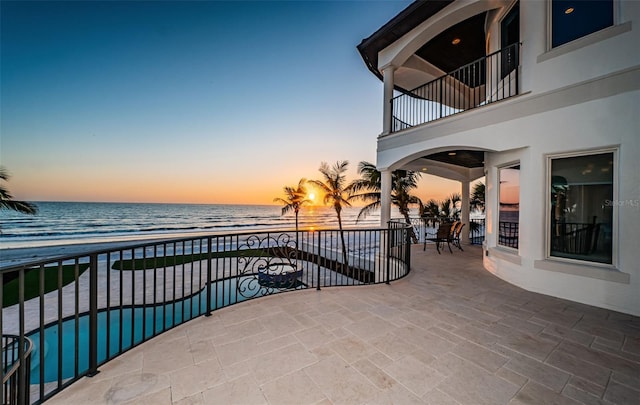 patio terrace at dusk with a balcony, a water view, and a view of the beach