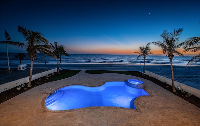 pool at dusk featuring a view of the beach, an in ground hot tub, a water view, and a patio