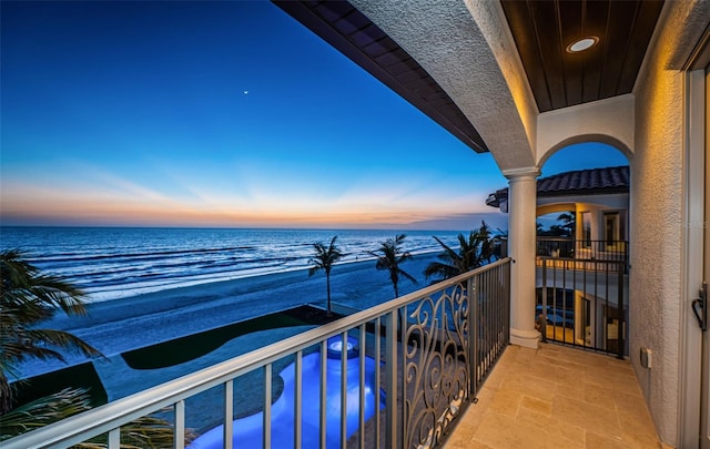 balcony at dusk featuring a water view and a beach view