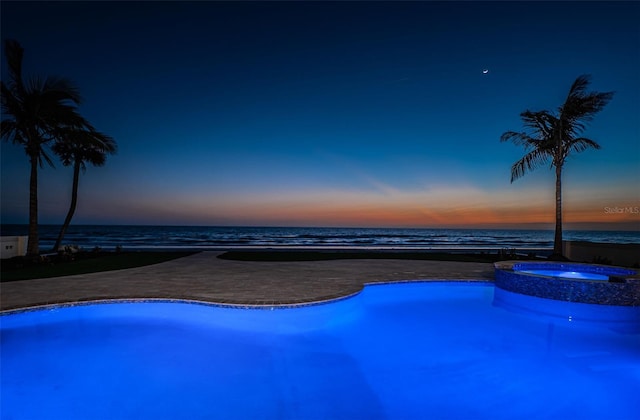 pool at dusk with an in ground hot tub, a beach view, and a water view