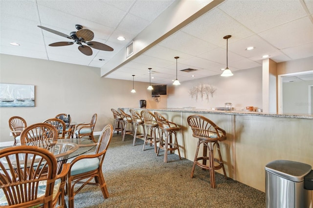 carpeted dining space featuring a paneled ceiling, ceiling fan, and indoor bar