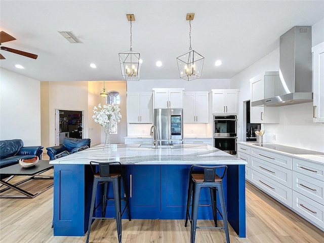 kitchen with white cabinetry, hanging light fixtures, a large island with sink, stainless steel appliances, and wall chimney range hood