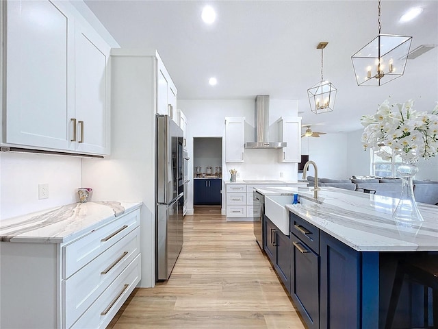 kitchen featuring wall chimney range hood, sink, white cabinetry, hanging light fixtures, and light stone countertops