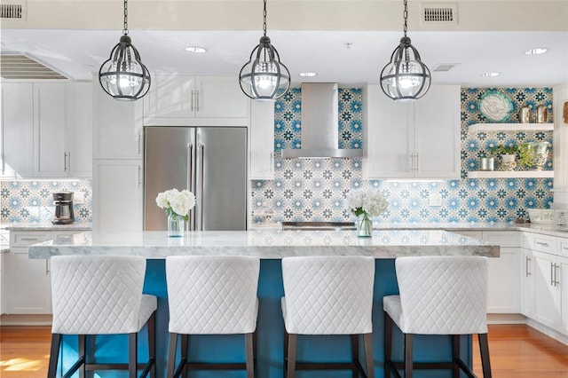 kitchen with a breakfast bar, white cabinetry, built in refrigerator, light stone counters, and wall chimney range hood