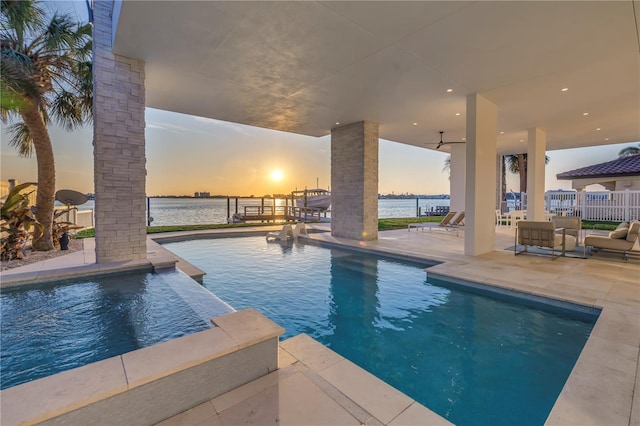 pool at dusk with a patio area, ceiling fan, and a water view