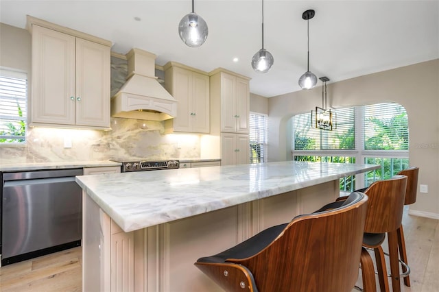 kitchen with pendant lighting, appliances with stainless steel finishes, cream cabinets, light stone countertops, and custom exhaust hood