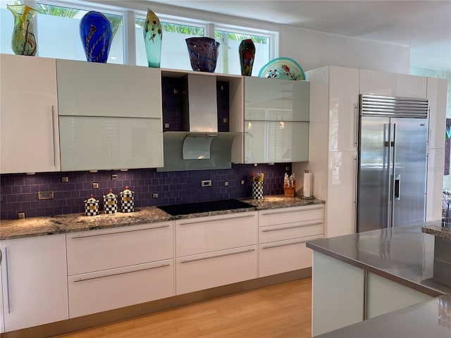 kitchen featuring black electric cooktop, backsplash, built in fridge, white cabinets, and wall chimney range hood