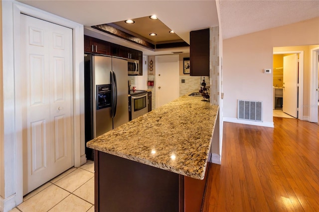 kitchen with appliances with stainless steel finishes, decorative backsplash, light stone counters, dark brown cabinets, and light wood-type flooring