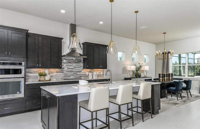 kitchen featuring a breakfast bar area, a kitchen island with sink, hanging light fixtures, decorative backsplash, and stainless steel double oven