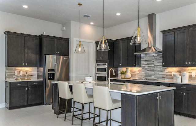 kitchen with sink, appliances with stainless steel finishes, a kitchen island with sink, decorative light fixtures, and wall chimney exhaust hood