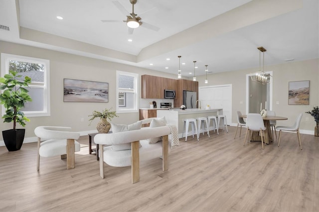 living room with ceiling fan with notable chandelier, a raised ceiling, and light wood-type flooring