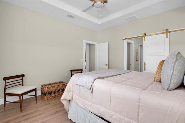 bedroom featuring a tray ceiling, light hardwood / wood-style floors, a barn door, and ceiling fan