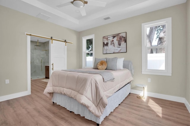 bedroom featuring a raised ceiling, a barn door, ensuite bath, and light hardwood / wood-style flooring