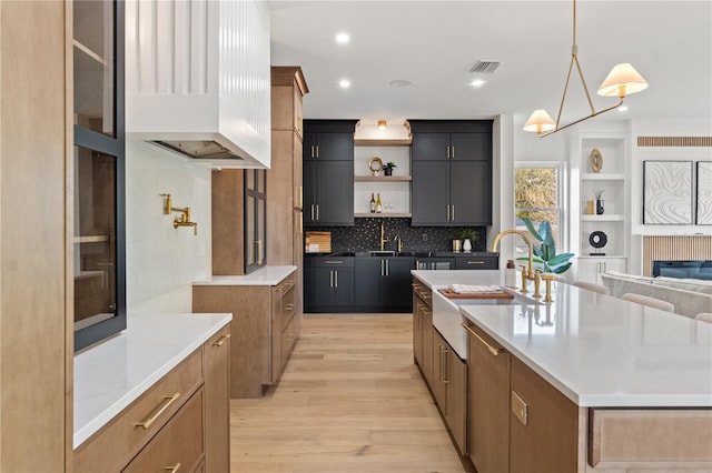 kitchen with open shelves, visible vents, light wood-style floors, a kitchen island with sink, and a sink