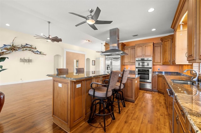 kitchen featuring sink, island range hood, appliances with stainless steel finishes, a kitchen island, and dark stone counters