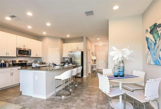 kitchen featuring appliances with stainless steel finishes, dark stone countertops, white cabinets, a center island with sink, and tile patterned floors