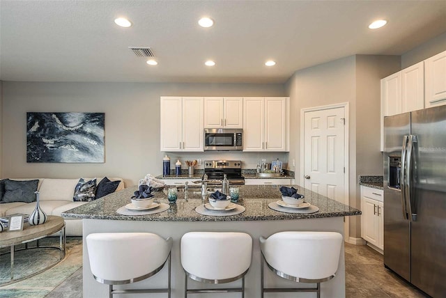 kitchen featuring an island with sink, white cabinetry, a kitchen bar, dark stone counters, and stainless steel appliances