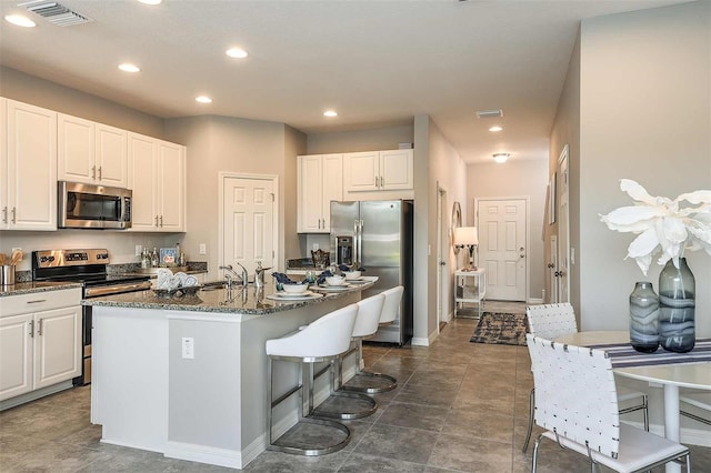 kitchen with white cabinetry, appliances with stainless steel finishes, a kitchen breakfast bar, an island with sink, and dark stone counters