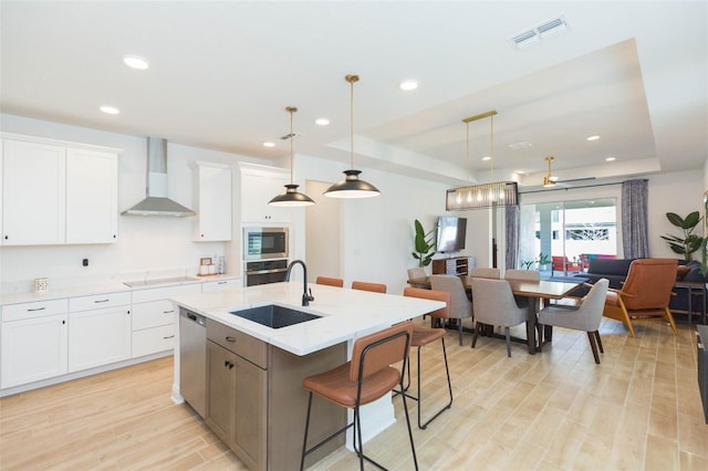 kitchen with wall chimney range hood, sink, stainless steel appliances, white cabinets, and a center island with sink