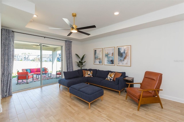 living room with a raised ceiling, ceiling fan, and light wood-type flooring