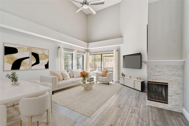 living room with ceiling fan, a stone fireplace, light hardwood / wood-style floors, and a high ceiling