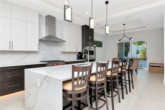 kitchen with an island with sink, a tray ceiling, a kitchen breakfast bar, wall chimney exhaust hood, and pendant lighting