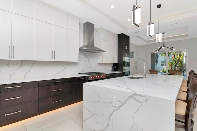 kitchen with a center island with sink, pendant lighting, a tray ceiling, a kitchen breakfast bar, and wall chimney range hood