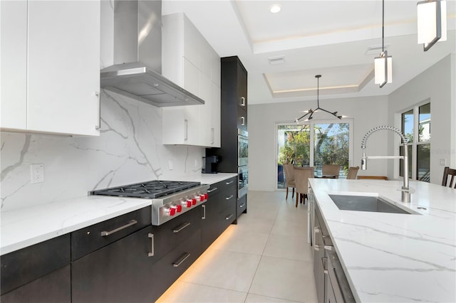 kitchen with wall chimney range hood, appliances with stainless steel finishes, a tray ceiling, white cabinets, and decorative light fixtures