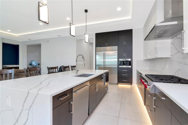 kitchen featuring hanging light fixtures, a tray ceiling, an island with sink, stainless steel appliances, and wall chimney range hood