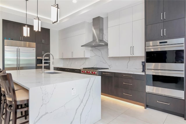 kitchen with wall chimney range hood, sink, appliances with stainless steel finishes, white cabinetry, and hanging light fixtures