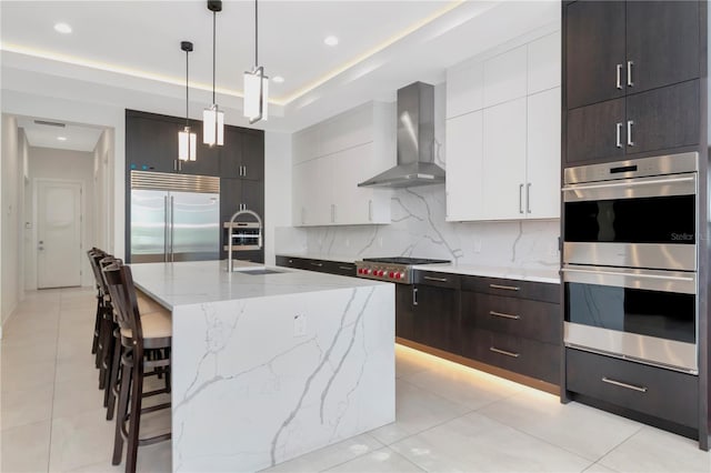 kitchen featuring appliances with stainless steel finishes, white cabinets, wall chimney range hood, hanging light fixtures, and a center island with sink