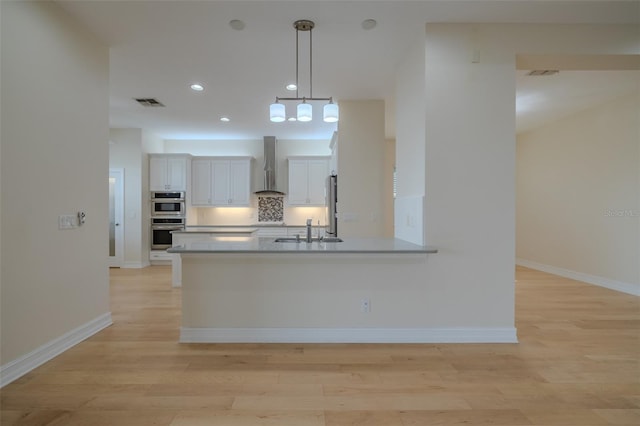 kitchen with a sink, wall chimney range hood, decorative backsplash, light wood finished floors, and decorative light fixtures