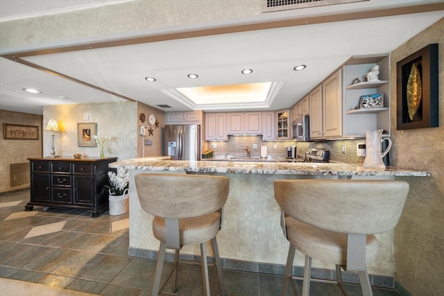 kitchen with light brown cabinetry, a raised ceiling, decorative backsplash, kitchen peninsula, and stainless steel appliances