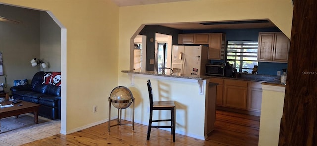 kitchen with a breakfast bar area, light wood-type flooring, kitchen peninsula, dark stone counters, and white refrigerator with ice dispenser