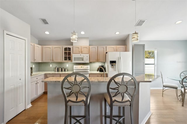 kitchen with hanging light fixtures, light brown cabinets, an island with sink, white appliances, and light stone countertops