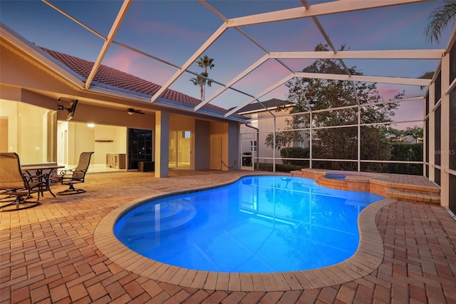 pool at dusk with an in ground hot tub, ceiling fan, a patio, and glass enclosure