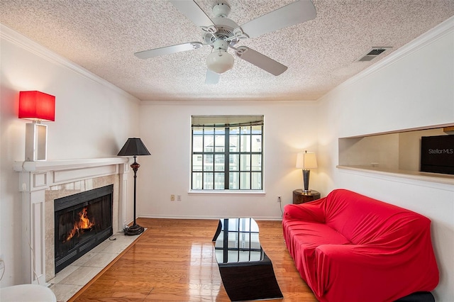 living room with crown molding, a textured ceiling, ceiling fan, a tiled fireplace, and light hardwood / wood-style floors