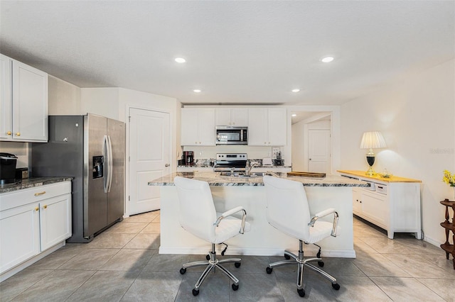 kitchen featuring light tile patterned floors, appliances with stainless steel finishes, a kitchen island with sink, light stone counters, and white cabinets