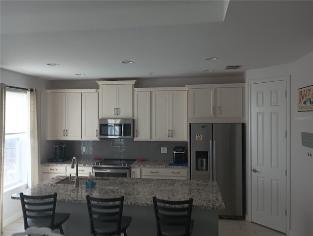 kitchen with a kitchen island with sink, white cabinetry, stainless steel appliances, light stone countertops, and a kitchen bar