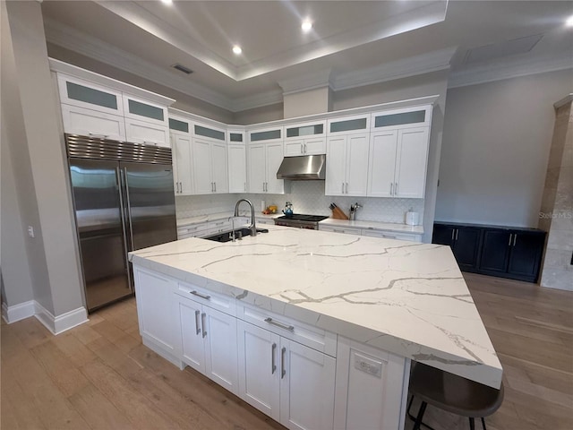 kitchen with a center island with sink, built in refrigerator, a tray ceiling, under cabinet range hood, and a sink