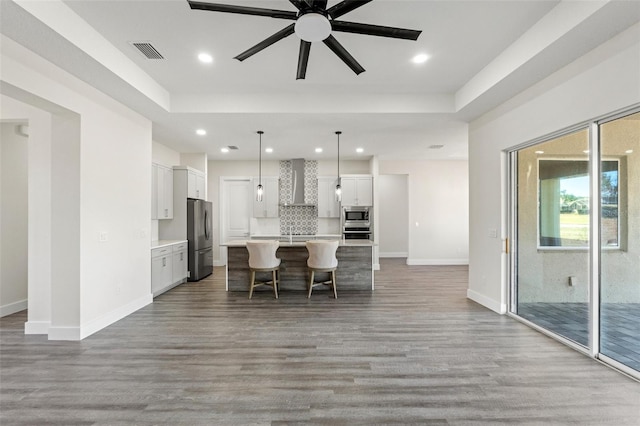 kitchen with wall chimney range hood, appliances with stainless steel finishes, white cabinetry, a center island with sink, and decorative light fixtures