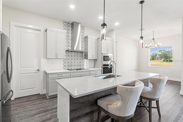 kitchen featuring stainless steel appliances, a center island with sink, wall chimney range hood, and decorative light fixtures