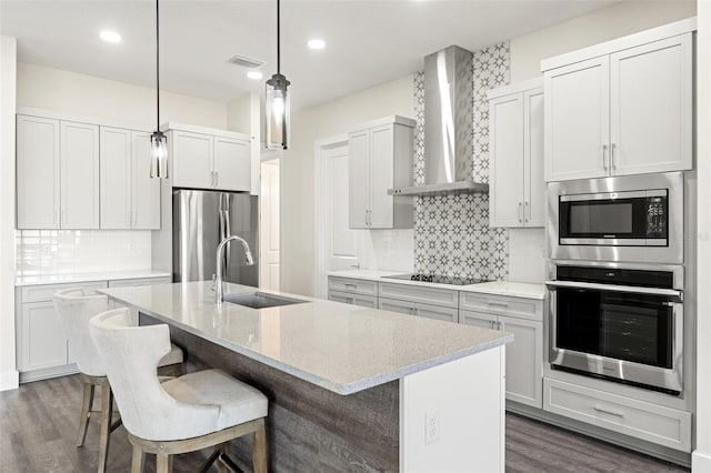 kitchen featuring wall chimney range hood, decorative light fixtures, an island with sink, and appliances with stainless steel finishes