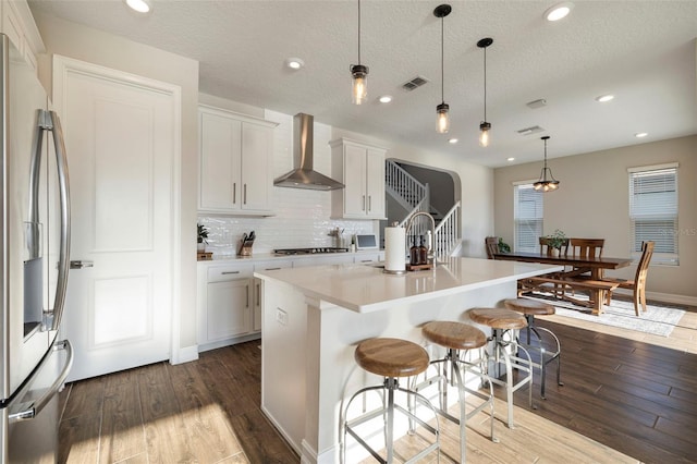 kitchen featuring wall chimney range hood, a kitchen island with sink, hanging light fixtures, white cabinets, and stainless steel fridge with ice dispenser