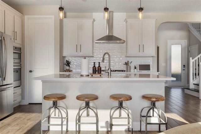 kitchen featuring white cabinetry, wall chimney exhaust hood, a kitchen island with sink, and hanging light fixtures