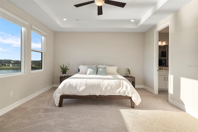 carpeted bedroom with a water view, ceiling fan, connected bathroom, and a tray ceiling